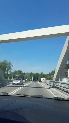The video appears to be captured from the perspective of someone driving a vehicle. The road ahead is paved and appears to be a two-lane road with a white line separating the lanes. There are guardrails on both sides of the road. On the left side of the road, there are trees and some buildings that resemble houses. On the right side of the road, there are also trees and houses with sloped roofs, suggesting a mountainous or hilly terrain. The sky is clear and blue, indicating a sunny day. The overall environment suggests a scenic drive through a rural or suburban area.