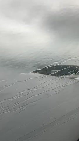 The video shows a view from an airplane window with raindrops running down the glass. Below, a landscape is visible through the smudged window, including what appears to be the wing of the airplane, water, and land with some structures. The sky is overcast and gray. The camera is stationary and the view is slightly distorted due to the raindrops and the window.