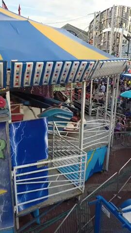 This media file captures a scene at an outdoor carnival or amusement park. The primary subject is a large, blue and yellow striped ride structure, likely a carnival ride, with visible mechanical elements like metal beams and cables. The ride features a blue facade decorated with white lightning bolt graphics and a series of lights. In the background, other carnival attractions, food stands with colorful signage, and a Ferris wheel are visible. Numerous people, including children and adults, are scattered throughout the scene, suggesting a lively atmosphere. Some are queuing for rides or browsing stalls, while others are observing the attractions.

The activity taking place is the operation of the carnival, with rides and vendors active. The time of day appears to be late afternoon or early evening, as the sky is a soft blue with hints of clouds, and the lighting suggests it's not yet fully dark. The weather seems pleasant, suitable for an outdoor event. Location cues include the presence of typical carnival setups and the distant structure of a Ferris wheel, indicative of a fair or amusement park. The overall mood conveyed is one of fun and recreation.

Several pieces of visible text can be identified. On the ride structure itself, the number "51" is prominently displayed. Another visible sign reads "THINK SAFETY FIRST," a safety reminder often found at such venues. The background signage for the food stands includes words like "CREPE," "CANDY," and "BASKETBALL," indicating the types of offerings at the carnival. The presence of these elements paints a clear picture of a bustling public gathering centered around entertainment and food.
