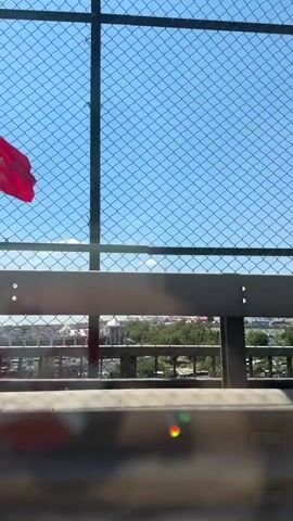 The video shows a view from inside a vehicle moving along a road. A large red flag with a white crescent moon and star, likely the Turkish flag, is prominently displayed and waving in the wind, seen through a chain-link fence. The fence appears to be part of a barrier along an elevated roadway or bridge. Beyond the fence, there are buildings and trees in the distance, suggesting an urban or suburban environment. The sky is clear and blue. The camera is angled downwards and towards the right, with the dashboard and part of the car's interior visible in the foreground. The movement of the vehicle is evident from the slight blur of the distant landscape and the perspective shift.