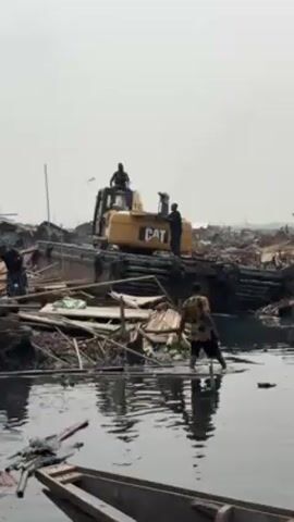 The video depicts a scene of destruction with a CAT excavator atop debris amid water. Several individuals are present: one is operating the excavator, another stands on top of the rubble near the excavator, one person wades in the water, and a fourth stands on the debris to the left of the excavator. The environment appears to be a slum or shantytown located on or near water, with debris scattered everywhere. The overall tone suggests a demolition or cleanup operation in progress.