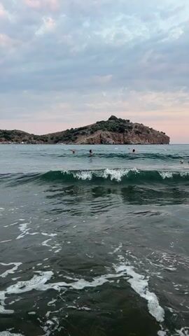 The video begins with a wide shot of a beach scene. In the foreground, waves crash onto the shore, creating white foam. The water is a deep blue-green. In the mid-ground, there are several people swimming or wading in the water. Some appear to be wearing swim caps. The sea stretches out to the horizon, where it meets a hazy sky with pale pink and grey clouds. In the distance, a rocky, tree-covered island rises from the water. As the video progresses, the camera moves closer to the water's edge. The waves become more prominent, and the foam covers the sand. The final shot is a close-up of a person's bare feet standing in the shallow water, with the white foam of a breaking wave washing over them. The overall mood is serene and natural.