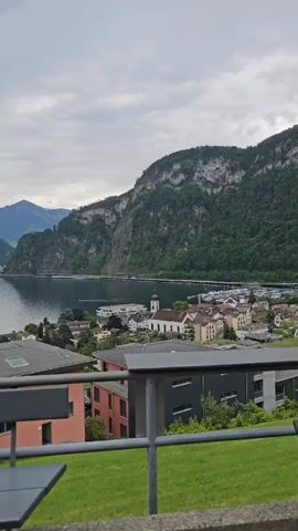 Here is a concise description of the video:

The video presents a scenic view of a town nestled beside a lake, framed by mountainous terrain. The shot is taken from an elevated position, likely a balcony or terrace, evident by the railing and structural elements in the foreground. The town features various buildings, with a notable church steeple rising among them. The lake stretches out, reflecting the cloudy sky above. A bridge or causeway is visible in the distance, connecting different parts of the shoreline. The mountains are covered in lush greenery, with rocky outcrops punctuating their slopes. The overall environment appears tranquil and picturesque.