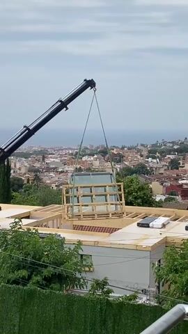 A crane is lifting a large glass structure on a wooden pallet over the rooftop of a building under construction. A person stands on the rooftop. In the background, a city skyline is visible.