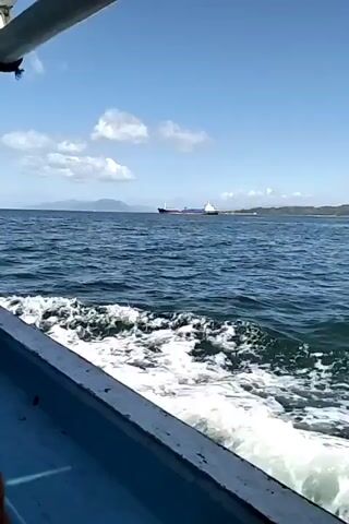 The video shows a view from a boat moving through the water. The water is choppy, with white foam trailing behind the boat, indicating movement. In the distance, a large cargo ship is visible on the horizon. The sky is blue with some scattered white clouds. Landmasses, possibly islands or a coastline, are visible in the far distance behind the cargo ship. The perspective is from the side of the boat, showing its blue hull. The sun appears to be shining, creating a bright environment.