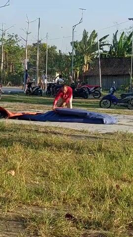 A person in a red shirt and beige pants is kneeling on a blue tarp on the ground outdoors. They are appearing to fold or manipulate the tarp with their hands. The tarp is elongated and appears to be made of a thick material. In the background, there are several motorcycles parked. Beyond the motorcycles, there is a line of trees and foliage, some of which are banana trees. There are also structures with thatched roofs visible. In the further background, there are tall poles with netting stretched between them, possibly for a sports field. The ground in the foreground is covered in grass. The sky is clear and blue, indicating a daytime scene.