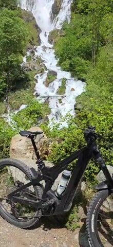 A black mountain bike is parked in front of a cascading waterfall. The waterfall is surrounded by lush green vegetation. The water is white and foamy as it rushes down the rocks. The bike has a water bottle in its cage. Two people are visible in the waterfall.