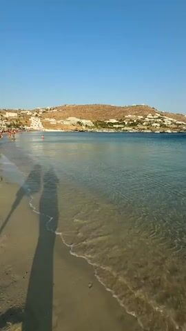This is a vertical video clip of a tranquil beach scene. The foreground features a sandy beach with gentle waves lapping at the shore. The water is crystal clear, revealing the sandy bottom beneath. Two prominent shadows of people walking are cast on the wet sand in the left portion of the frame. In the mid-ground, the clear blue sea stretches out towards the horizon.

The background showcases a picturesque coastal landscape. A hillside dotted with white buildings, characteristic of Mediterranean architecture, rises from the coast. Several boats, including sailboats, are anchored in the bay, adding to the serene atmosphere. The sky above is a clear, vibrant blue, suggesting fair weather and a sunny day.

The time of day appears to be late afternoon or early evening, given the soft, elongated shadows. The overall mood is peaceful and idyllic, evoking a sense of relaxation and vacation. The location is identified as Agios Ioannis Diakoftis, Greece, which aligns with the visual cues of the landscape and architecture. There is no visible text in the video clip.
