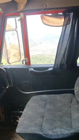 The video shows the interior of a truck cab, looking out the passenger side window. The seat is upholstered in a dark grey speckled fabric. The door panel is black plastic with a dark grey handle. Outside the window, a landscape of rolling hills and green trees is visible under a bright, overcast sky, suggesting daytime. The terrain appears somewhat arid with patches of vegetation and possibly some dry riverbeds or valleys. The truck is likely on a road or path in a rural, possibly mountainous, area, given the landscape. There is no text visible in the media. The perspective suggests the truck is moving, as the view is slightly jiggling.