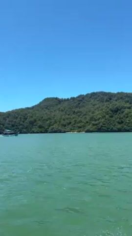 The video shows a sunny day on the water. The water is a turquoise color, and the sky is clear blue. There is a green, tree-covered hill in the background. A tour boat is travelling on the water. White water splashes behind the boat in the foreground. Another boat is also visible in the distance.
