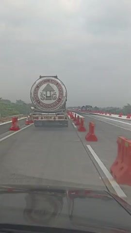The video shows a view from the driver's seat of a vehicle moving on a road under construction. The road has construction barriers and cones lining the lanes. In front of the vehicle, a large tanker truck with the logo SLG INDONESIA is visible. The sky is overcast, and the surrounding area appears to be rural with trees and vegetation. The truck is driving in the same direction as the vehicle, and other vehicles can be seen in the distance.