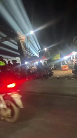 The video shows a nighttime street scene with several people and motorcycles. Bright lights illuminate the area, suggesting a market or gathering. Some individuals are gathered around motorcycles, while others are near food stalls or vendors with tents. The overall atmosphere appears casual and social.