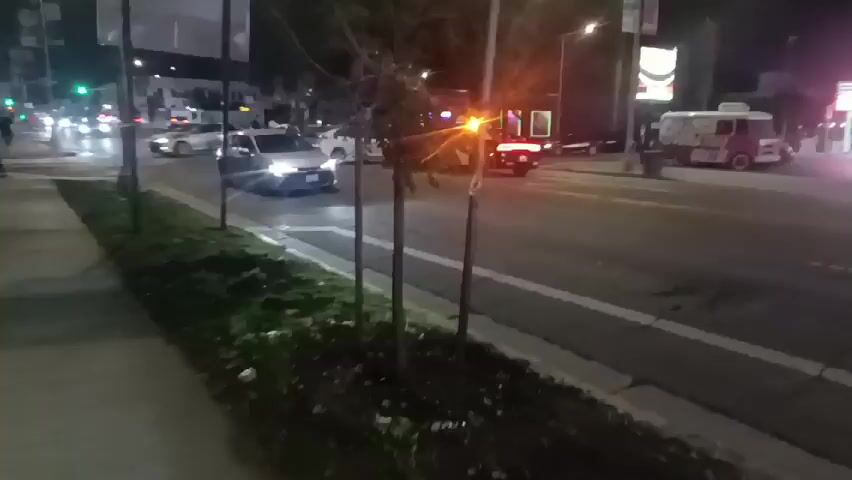 The video, taken from a sidewalk at night, shows a police car parked on a city street. Its emergency lights are flashing. Two white cars are also present, parked nearby.  Several people are visible near the vehicles, though their actions are unclear. The street is illuminated by streetlights, and other cars pass by in the background. The sidewalk is bordered by a strip of grass and small trees.
