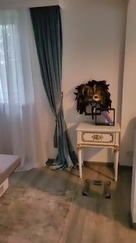 The video pans across a bedroom. On the left, a white sheer curtain is visible, partially covered by a teal curtain. A rug lies on the wooden floor. Next to the curtain is a small nightstand with a mask on top and a tablet. To the right of the nightstand, a digital scale is on the floor. The camera then moves to the right, showing a bed with a grey mattress on a pallet base, covered with a patterned gold blanket and several decorative pillows. A large wall art depicting a hallway with mirrors is above the bed's headboard.