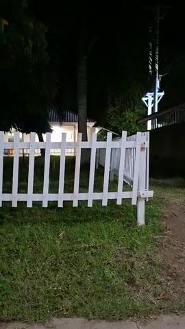 The video shows a nighttime scene in Tambonbaroh, Indonesia. A white picket fence runs along the foreground, with lush green grass behind it. Beyond the fence, several trees with dark foliage are visible, their trunks illuminated by faint lights from a building in the distance. The building appears to have large windows that emit a warm, yellowish glow. The overall atmosphere is tranquil and peaceful.

In the far distance, a single person can be seen sitting down, adding a touch of human presence to the serene environment. The time of day is clearly night, indicated by the darkness of the sky and the artificial lighting. There are no specific weather cues, but the grass appears healthy, suggesting it's not a dry season. The location cues are limited to the mention of Tambonbaroh, Indonesia. No text is visible in the video. The mood evoked by the scene is one of calm and quiet contemplation, enhanced by the soft lighting and the stillness of the night.