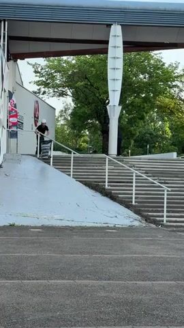 The video captures an outdoor scene featuring a person attempting a scooter trick down a handrail. The setting includes a large concrete staircase with white handrails on both sides, and a sloped concrete ramp adjacent to the stairs on the left. In the background, there's a white building with some red graphics and partially visible text (including "TEAM") and a logo. Beyond the building, lush green trees are visible, and a distinctive tall, white, multi-faceted pillar stands to the right of the ramp.

The main subject, wearing a white helmet, black t-shirt, and dark pants, rides a scooter towards the handrail at the top of the ramp. They successfully jump onto the handrail, performing a grind. However, upon reaching the end of the rail, they lose balance and fall off the scooter, which slides forward. The rider remains standing after the fall. Another person, dressed in dark clothing, is standing on the stairs further back, observing or possibly filming the attempt.

The scene takes place during the day, under clear and bright conditions. The overall atmosphere suggests a public park or recreational area in Saint-Priest, France. A parked bicycle is briefly visible further up the stairs after the scooter rider falls.