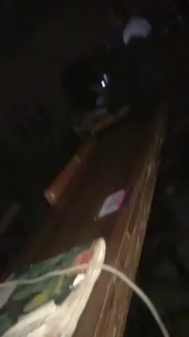 The camera pans upward over a dark wood surface, revealing several objects including a brown-handled hammer, a small container, and a bottle of water.  A person wearing a pink shirt is visible at the bottom of the frame. The room is dark and sparsely lit. The camera movement is shaky.
