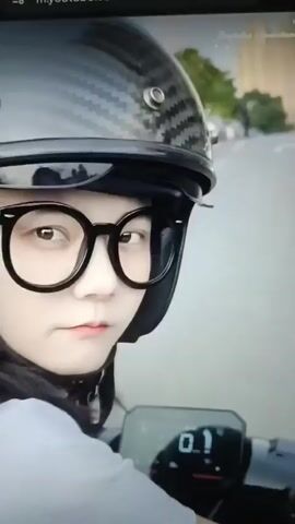 This is a close-up video of a person wearing a motorcycle helmet and glasses. The helmet is black with a carbon fiber pattern and some graphics on the side. The person is looking at the camera and then turns their head to the side. The video shows a dashboard on a motorcycle or scooter with a digital display. The background appears to be an outdoor scene with trees and cars. The person is wearing a white shirt. The video is short and seems to be a clip from a larger video or social media post.