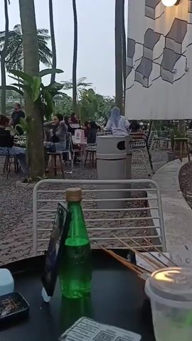 The video shows an outdoor cafe with several people sitting at tables. Tall palm trees are visible in the background, and the ground is covered in gravel. In the foreground, a table holds a green bottle of soda, a smartphone, and some skewers. Several patrons are seated at tables, some with drinks and food. The overall atmosphere appears relaxed and casual.