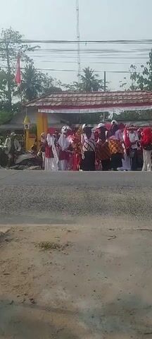 A motorcycle passes on the left side of the screen, blurring as it moves. Ahead, a group of people are gathered. Some are dressed in traditional Indonesian clothing with red and white accents, suggesting a festive occasion or a national holiday. A young person in the center wears a gold headdress and a red costume, possibly participating in a parade or performance. Others are observing the proceedings. The setting appears to be outdoors, possibly a street or a public gathering area, with trees and a building structure in the background. The overall atmosphere seems celebratory.