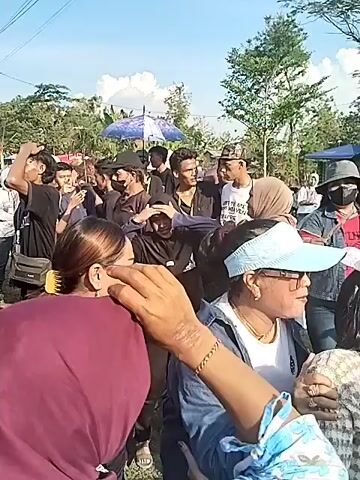 A medium shot shows a crowd of people, mostly looking in the same direction. Many are wearing head coverings, including hijabs and baseball caps. A man in a black t-shirt smiles slightly at the camera. In the background there are trees, blue umbrellas and a vehicle. The scene appears to be outdoors in daylight. There is music playing in the background.