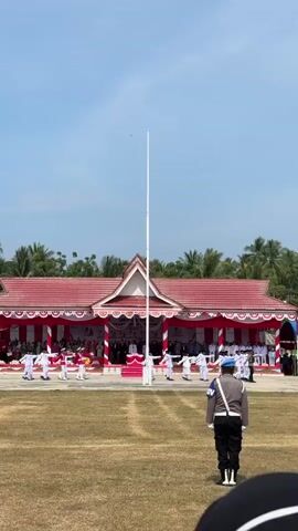 A police officer in a blue shirt and black pants stands in the foreground facing away from the camera, looking towards a large open field with a flagpole in the center. In the background, a group of people in white uniforms are performing a synchronized dance or drill in front of a red and white building with a white roof. There are spectators seated in the background. The sky is clear and blue, and palm trees are visible on the horizon.