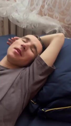 This is a short video of a young man lying on a dark blue surface, possibly a bed or sofa, with his head resting on his arm. He is wearing a dark grey t-shirt. The man's eyes are closed, and he appears to be either sleeping or resting. His facial expression suggests a state of relaxation, though at one point he wrinkles his nose and makes a slight grimace. The background shows a striped wall and a sheer white curtain with an ornate pattern, suggesting an indoor setting. The lighting is soft and appears to be artificial, indicating it could be daytime or nighttime. There is no visible text in the video. The overall mood is one of peaceful repose.