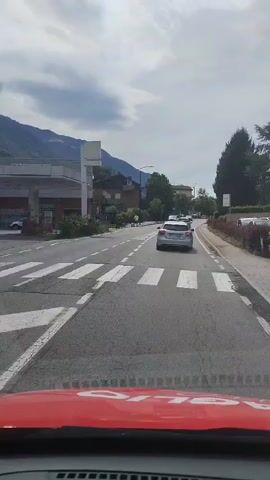 The video is taken from inside a vehicle, looking forward. The vehicle is on a paved road with a pedestrian crossing marked in white stripes in the foreground. There are two lanes of traffic. Ahead, a white car is in the right lane, and a gray car is in the left lane. Further down the road, there is a traffic light and more cars. On the left side of the road, there are buildings and trees. On the right side of the road, there is a sidewalk with some bushes and trees. The sky is partly cloudy.