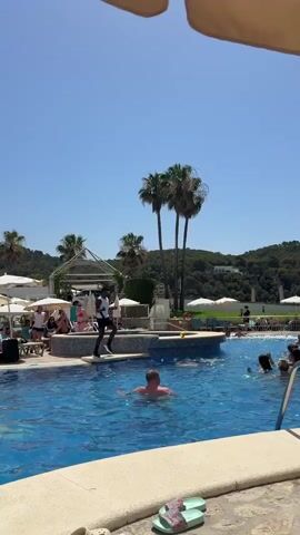 The video captures a lively scene at an outdoor swimming pool on a sunny day. The bright blue sky and clear weather suggest it's midday. Palm trees and lush green hills in the background indicate a warm, tropical or Mediterranean setting, consistent with the specified location of Es Camp De Mar, Spain.

The primary subject is a large group of people enjoying the pool. Many are submerged in the water, some interacting with each other, while others seem to be participating in an organized activity. On a raised platform at the edge of the pool, a man dressed in a white shirt and dark shorts is actively leading the group, likely an instructor or entertainer. His posture and the attention of the people in the pool suggest he is conducting an exercise class or some form of pool aerobics.

A few individuals are visible lounging or standing around the pool area, some under umbrellas, indicating a resort or leisure facility. The water is a vibrant blue, and the edge of the pool is lined with light-colored tiles and a stone-like pathway. A metal pool ladder is prominently featured in the foreground.

The overall mood appears to be cheerful and energetic, with people actively engaged in a recreational activity under a clear, sunny sky. There is no visible text within the frame.