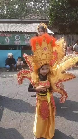 A young girl in a colorful, ornate costume with a large headdress and wing-like appendages stands in the foreground of a street procession. She is wearing a yellow and red outfit with intricate gold detailing. Behind her, an adult woman also in costume, likely a supervisor or participant, stands with her hands clasped. Other participants, including children and adults in various traditional and festive attire, are visible further down the street, moving in a procession. Spectators are seated or standing along the side of the road, observing the event. The environment appears to be an outdoor public space with buildings and trees lining the street. The overall scene depicts a cultural celebration or parade.