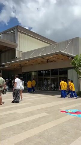A group of at least seven children, all wearing bright yellow t-shirts and blue pants, walk in single file past a storefront with a sign that reads "PREM." The children are walking from left to right across the frame. They are on a paved outdoor walkway. To the left of the children, two adults, one wearing a white t-shirt and patterned pants and the other wearing a light-colored shirt and skirt, are walking in the opposite direction. Behind the children and the store is a building with a light-colored facade and a dark-colored roof structure. Above the store's entrance is a light-colored awning with a woven texture. The sky above is blue with scattered white clouds.