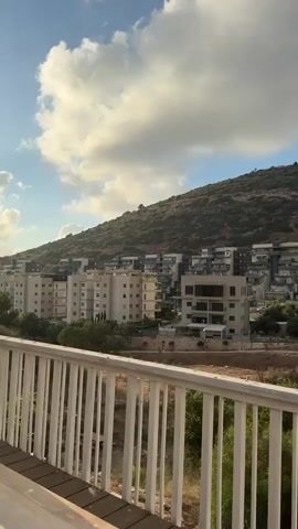 The video shows a view from a balcony or deck, looking out at a hillside with buildings and trees. In the foreground, there is a white railing and wooden deck. The hillside is covered in vegetation and features several multi-story apartment buildings. The sky above is blue with scattered white clouds. The lighting suggests it is daytime, possibly late afternoon or early morning due to the angle of the sun. There are no people or discernible actions taking place in the video.