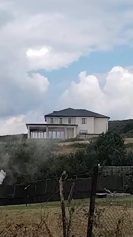The video shows a modern, two-story house with a dark roof, situated on a hill overlooking a wooded area. The house has large windows and a glass-enclosed extension. In the foreground, there is a chain-link fence and some dry vegetation. A plume of smoke is visible rising from behind the fence on the left side of the frame. The sky is cloudy with patches of blue.