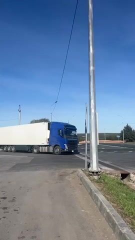 The camera remains still in a medium shot of a blue and white semi-truck as it waits on an asphalt road to turn onto a road perpendicular to the viewer, which is visible behind it. There is a grassy verge with a storm drain to the camera's right, beside a metal pole on a concrete and asphalt base. The sky is blue, and there are trees and electrical lines in the background.