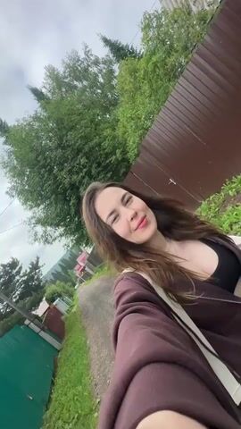 A young woman with long brown hair is taking a selfie outdoors. She is wearing a brown zip-up hoodie over a black top. Her eyes are closed and she has a slight smile on her face. The background appears to be a rural or suburban setting, with a gravel road, a brown wooden fence, green grass, and trees. The sky is overcast. The video is shot from a low angle, with the woman's arm holding the phone visible in the foreground.
