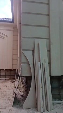 The video shows a construction site with a building facade under renovation. The facade is made of light-colored stone or plaster, with decorative elements like horizontal banding and a curved arch. Several pieces of building material, including long, flat planks and curved decorative pieces, are leaning against the wall. A metal ladder is also visible. The ground is dirt and debris. The sky is visible with some clouds, and power lines run across the background. The overall impression is of ongoing construction or repair work on the exterior of a building.