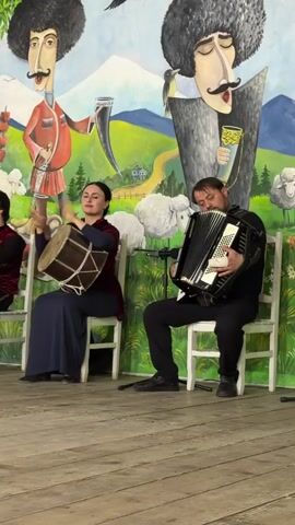 A WOMAN SITS ON A CHAIR TO THE LEFT, PLAYING A DARBUKA DRUM WITH HER HANDS. A MAN SITS ON A CHAIR TO HER RIGHT, PLAYING AN ACCORDION. BOTH ARE SEATED IN FRONT OF A MURAL DEPICTING TWO CARTOONISH FIGURES IN TRADITIONAL CLOTHING, WITH SHEEP AND LANDSCAPES IN THE BACKGROUND. THE WALL APPEARS TO BE DECORATED WITH FLOWER MOTIFS. THE FLOOR IS MADE OF WOODEN PLANKS.