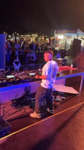 A male DJ stands behind a DJ booth on a stage at night. He wears a white t-shirt with a graphic print and dark pants. The DJ is operating the DJ equipment and appears to be performing for a crowd of people who are visible in the background. The crowd is standing behind a barricade. Two plumes of white smoke are rising from the DJ booth. Lights and a stage setup are visible behind the DJ.