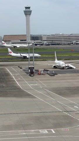The video shows an aerial view of an airport. Several airplanes are visible on the tarmac, including two Japan Airlines planes. One Japan Airlines plane is parked with a mobile stairway attached to its door. Another Japan Airlines plane is taxiing on a runway. In the background, a tall control tower stands prominently, and a long airport terminal building is visible. The sky is overcast.