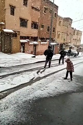 The video shows a street scene in what appears to be a city with buildings of similar height and architectural style lining the road. It is snowing, and a layer of snow covers the ground. The road is partially covered in snow and appears wet in places. There are a few people walking on the street, including two young children, one in a pink and black coat and the other in a blue coat. Behind the children, three adults walk away from the camera. A car is parked on the side of the street, also covered in snow. Overhead power lines are visible, and streetlights stand on the sidewalk. The weather is overcast and the overall lighting is dim due to the snowfall.
