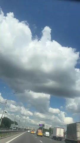A high-angle, vertical, handheld video shot captures a bright blue sky filled with large, fluffy white and gray clouds, suggesting a partly cloudy day. The camera appears to be inside a vehicle, likely moving forward on a highway. The perspective looks upward, capturing the vast expanse of the sky. Below the sky, a highway stretches into the distance, with several vehicles visible. Yellow taxis, white vans, and a large white truck with a closed trailer are among the traffic. On the left side of the highway, there's a concrete barrier and overhead lights. The right side of the highway is lined with trees and some buildings, including a tall white building in the background. The overall impression is a snapshot of a typical drive on a highway under a dramatic sky.