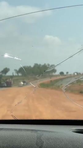 The video shows a view through a cracked windshield, revealing a dirt road leading to a paved road. Several vehicles, including a truck and a tanker, are seen driving on the paved road. Trees are visible in the background under a cloudy sky.