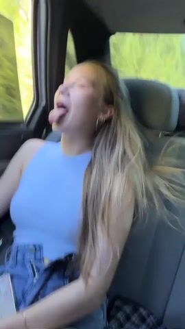 The video shows a young woman with blonde hair sitting in the passenger seat of a car. She is sticking her tongue out and her hair is blowing in the wind, suggesting the car window is down. She is wearing a blue tank top and denim shorts. The background outside the window shows greenery, indicating they are likely driving through a rural or wooded area. She is holding a phone in her left hand.