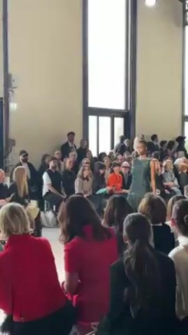 A model walks the runway at a fashion show in front of a seated audience. The model is wearing a floor-length, dark green gown with shimmering details. The audience members are varied in age and attire, many holding phones to record. The setting appears to be a bright, modern hall with tall windows. The atmosphere suggests a high-end fashion event.