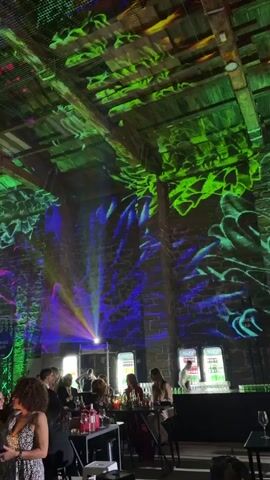 This is a video of an indoor event with people sitting at tables. The walls and ceiling are illuminated with colorful, abstract projections that shift and move. The projections include swirling patterns in green, blue, purple, and red, resembling smoke or liquid. There are exposed wooden beams on the ceiling, and the walls appear to be made of brick. Some people are visible sitting at tables with drinks, while others are standing or walking in the background. A bar area with illuminated shelving is visible on the right side of the frame. The overall atmosphere suggests a party or celebration with a light show.
