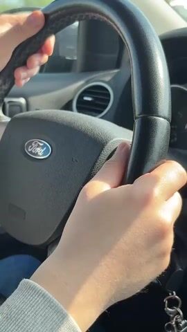 A close-up shot shows a pair of hands gripping the steering wheel of a Ford vehicle. The hands, belonging to a person wearing a gray sweater, are positioned at approximately the 10 and 2 o'clock positions. The Ford logo is prominently displayed on the center of the steering wheel. The car's interior, including the air vents and dashboard, is visible in the background. The lighting suggests that the scene is taking place during the day. There is music playing.