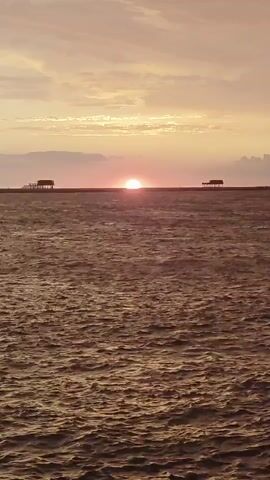 The video shows a sunset over a body of water. The sun is a bright orange orb partially obscured by a horizontal line, likely a distant shore or structure. Two small, dark, rectangular structures resembling small buildings or platforms are visible on this line, one on the left and one on the right of the sun. The water in the foreground is choppy and reflects the warm colors of the sky. The sky itself is filled with soft, diffused clouds in shades of pink, orange, and yellow. The overall mood is serene and tranquil.