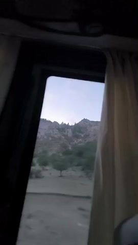 The video captures a view from inside a vehicle, looking out a window. The landscape outside is mountainous and appears somewhat arid, with sparse vegetation. In the distance, a transmission tower is visible on top of a rocky hill. The sky is a light blue with hints of pink, suggesting either dawn or dusk. The camera motion indicates the vehicle is moving, creating a blurred effect on the scenery. A portion of a beige curtain is visible on the right side of the frame, further confirming the perspective from within a vehicle. The overall scene is one of travel through a rugged, natural environment.