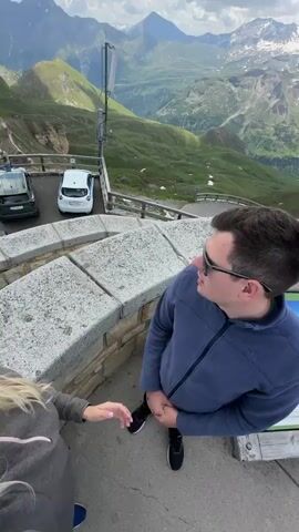 The video begins with a high-angle shot of a couple posing for a selfie on a stone observation deck. The woman, with blonde hair and sunglasses, smiles at the camera while holding it. Beside her, a man wearing sunglasses and a blue fleece jacket looks off to the side. In the background, there are several cars parked, and beyond them, a vast, lush green mountain range stretches out under a partly cloudy sky. The scene transitions to a slightly wider view showing more people on the observation deck. A woman in a purple jacket is gesturing with her phone, and other individuals are looking at informational displays. The camera then pans to show more of the observation deck and the surrounding mountainous landscape, emphasizing the elevation and scenic beauty.