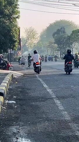 The video shows a street scene with several motorcycles and a bicycle in motion. The road appears to be asphalt with a dashed white line. On the left side of the road, there is a sidewalk with some trees and buildings. The sky is hazy, suggesting either early morning or a dusty atmosphere. People on motorcycles are wearing helmets and casual clothing. One person is riding a bicycle, carrying bags. The overall impression is that of a busy street during daylight hours, likely in an urban or suburban environment. There is some debris visible on the side of the road.